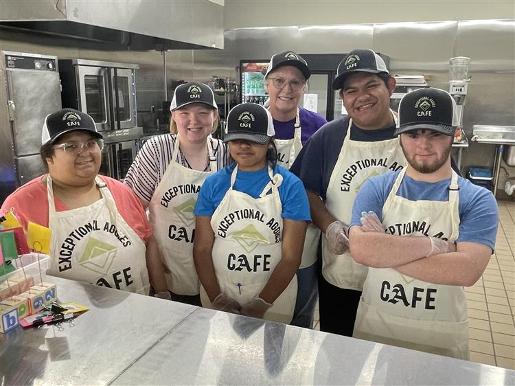 five students standing with their teacher. They are all wearing white aprons and black hats. They are standing in a kitchen.
